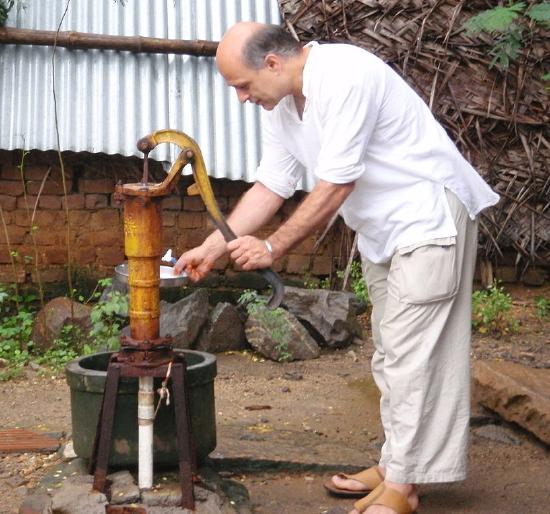 A man operating a hand pump with his left hand while his right hand is holding a basin to collect water.