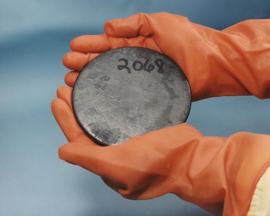 Round, flat billet of uranium with 2068 written on it in marker. It is held by a person wearing thick gloves.