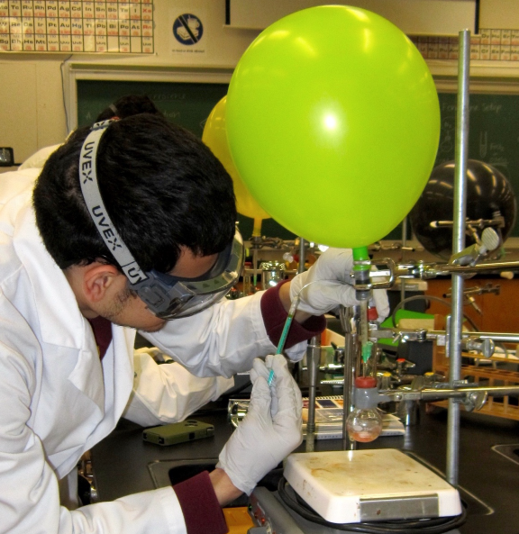   A lab worker leaning towards balloon running the Grignard reaction using a syringe to insert inert gases.