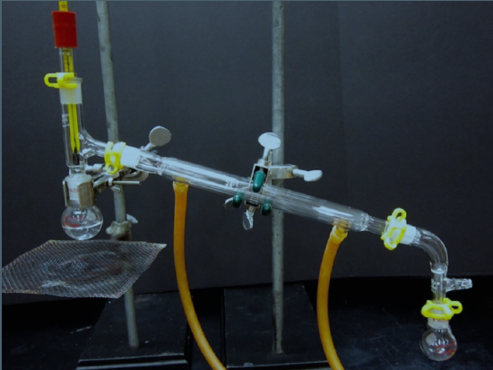  Distillation apparatus. A flask is connected to a vertical tube, with flows into a condenser, a tube that leads down and away from the vertical tube, ending in another flask. Two outlet hoses attach to the bottom of the condenser.