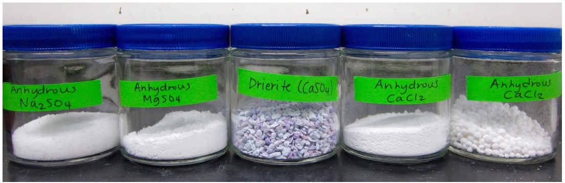   Set of five jars. From left to right: jar labelled "anhydrous N a 2 S O 4" filled with white powder. Jar labelled "anhydrous M g S O 4" filled with white powder. Jar labelled "drierite (C a S O 4)" filled with small pebbles of grey solid. Jar labelled "anhydrous C a C l 2" filled with white powder. Jar labelled "anhydrous C a C l 2" filled with small white spheres.