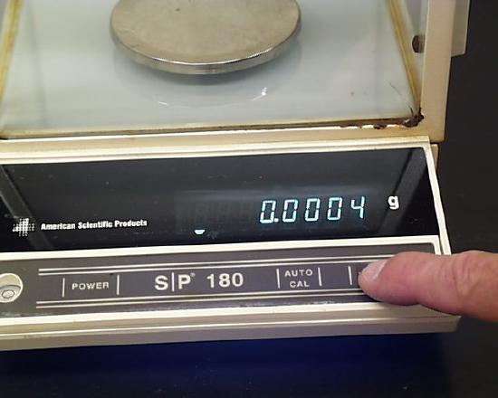 A hand is shown pressing the tare button on the balance. The balance now displays 0.0004 grams.