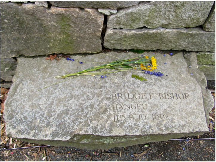 Gravestone with the words: Bridget Bishop. Hanged, June to 1692. Yellow flowers on top.