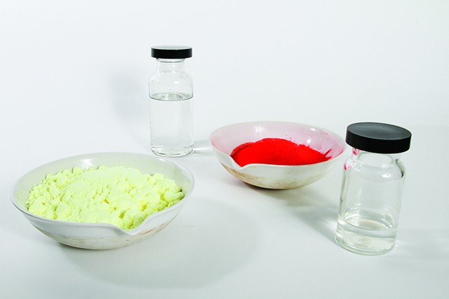 This photo shows two vials filled with a colorless liquid. It also shows two bowls: one filled with an off-white powder and one filled with a bright red powder.