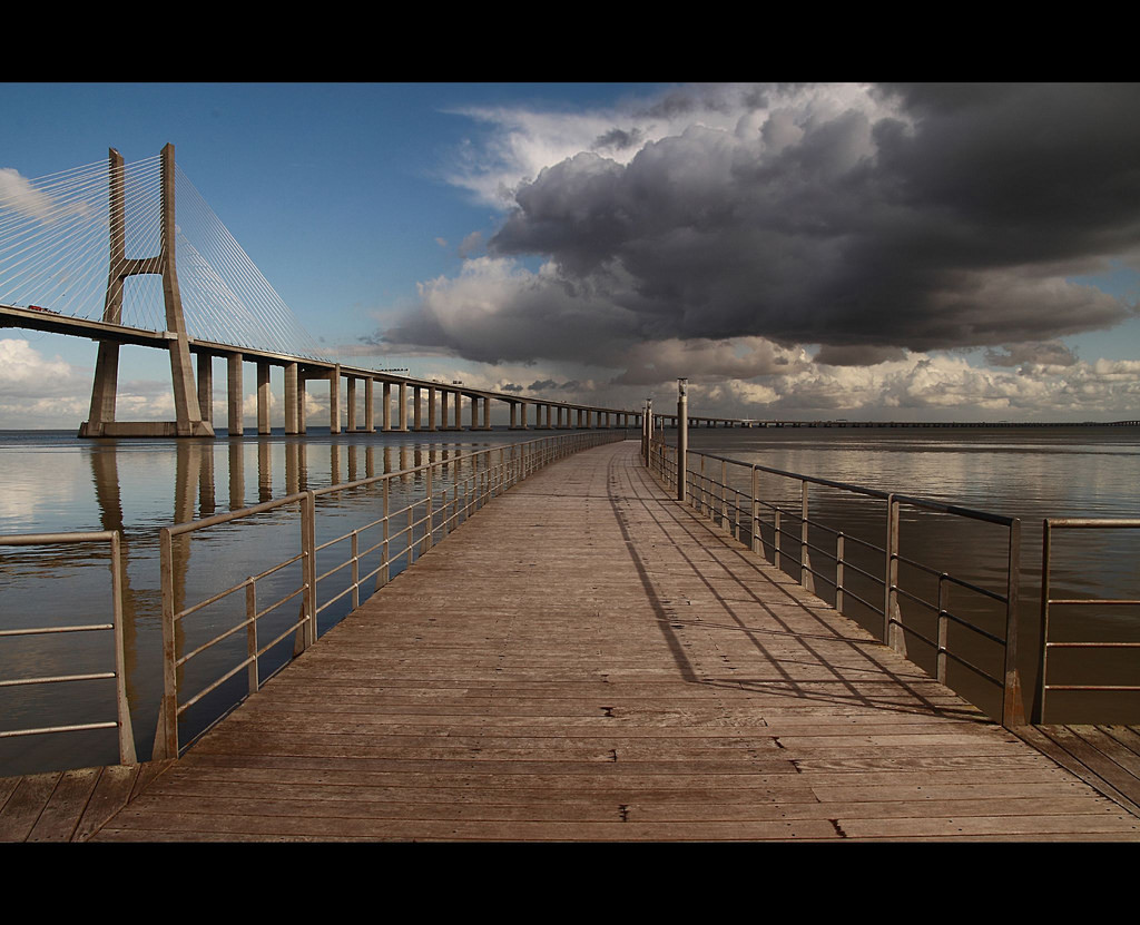Photo of a long wooden boardwalk that extends toward the horizon and disappears. In the background on the left is a graceful suspension bridge.