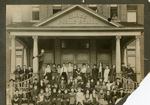 Vintage, sepia-toned photograph of roughly 50 children sitting on the steps in front of the Globe Public School.