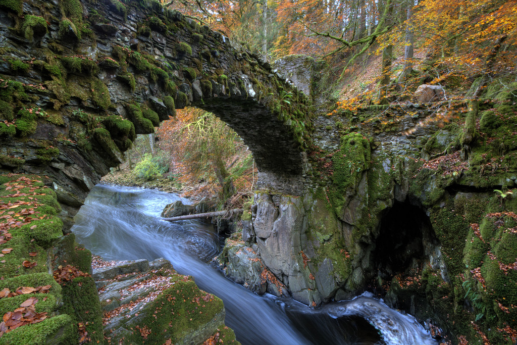 Photo of a rushing mountain stream beneath and old arched stone bridge. Mossy banks, brilliant orange leaves on the trees and ground.