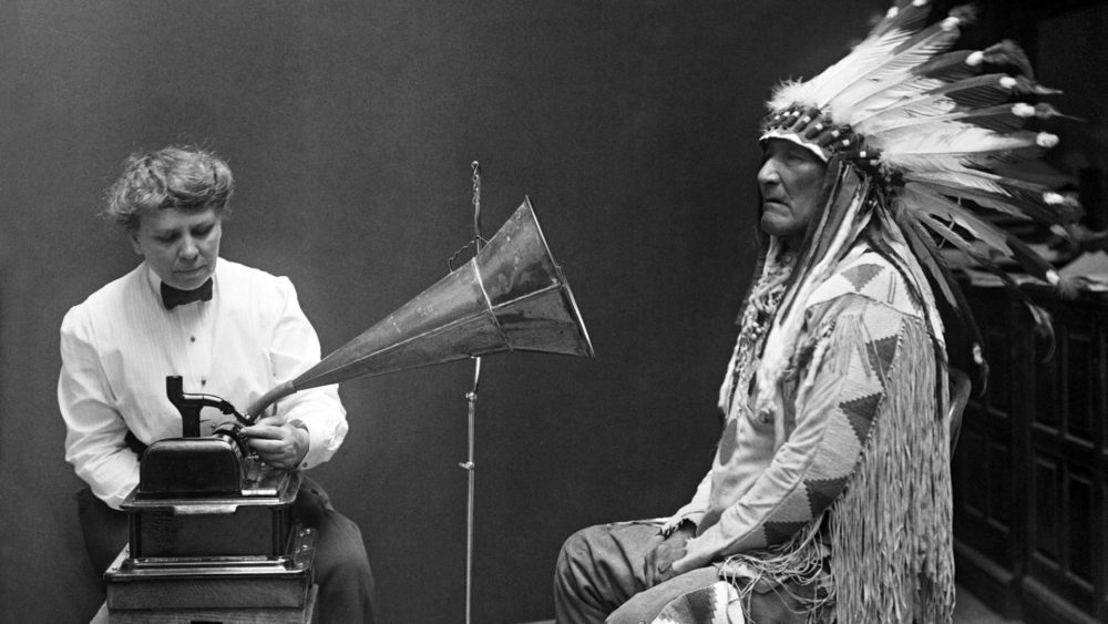 Frances Desmore sits with a megaphone in the face of Mountain Chief while he wears a traditional headress.