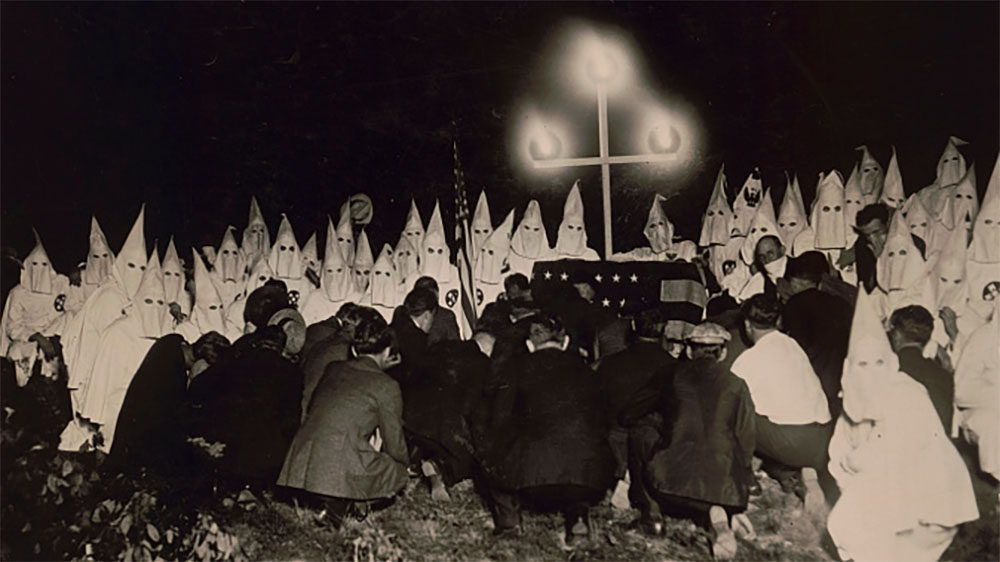 KKK gathering of a burning cross, 30-50 people in white robes and pointed white masks, and others in suits down on the ground.