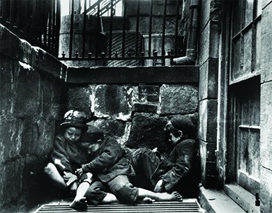 A photograph shows three small children, shabbily dressed and barefoot, asleep in a heap over a steam grate slightly below street level.