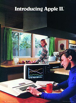 An advertisement shows a young man seated at a kitchen table working on a 1970s-style computer. A woman, who is preparing food at the kitchen counter, looks over her shoulder at him and smiles.