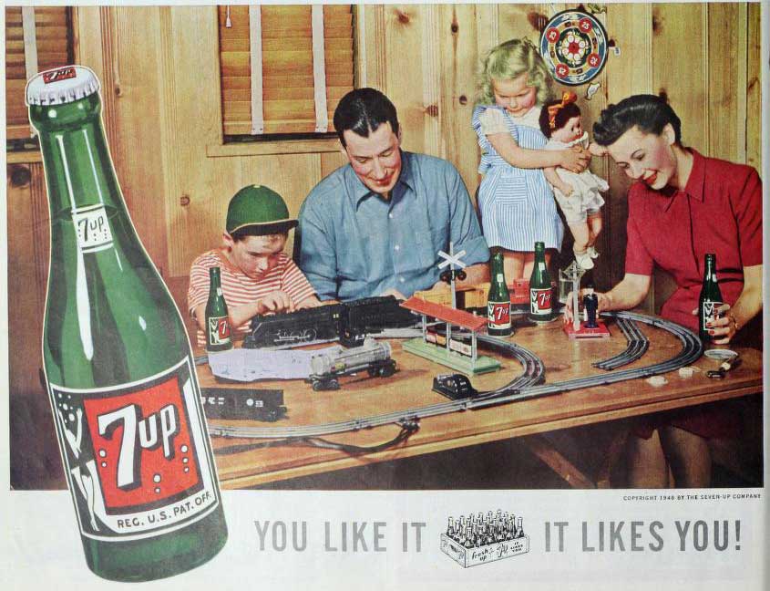 A 7-Up soda advertisement from the mid-twentieth century. The advertisement features a mother, father, son, and daughter playing with a table-top electrical train set and bonding over bottles of 7-Up soda. The advertisement features a large image of a bottle of 7-Up soda in the left margin and features the tagline â€˜You Like It; It Likes You!' at the bottom of the image.