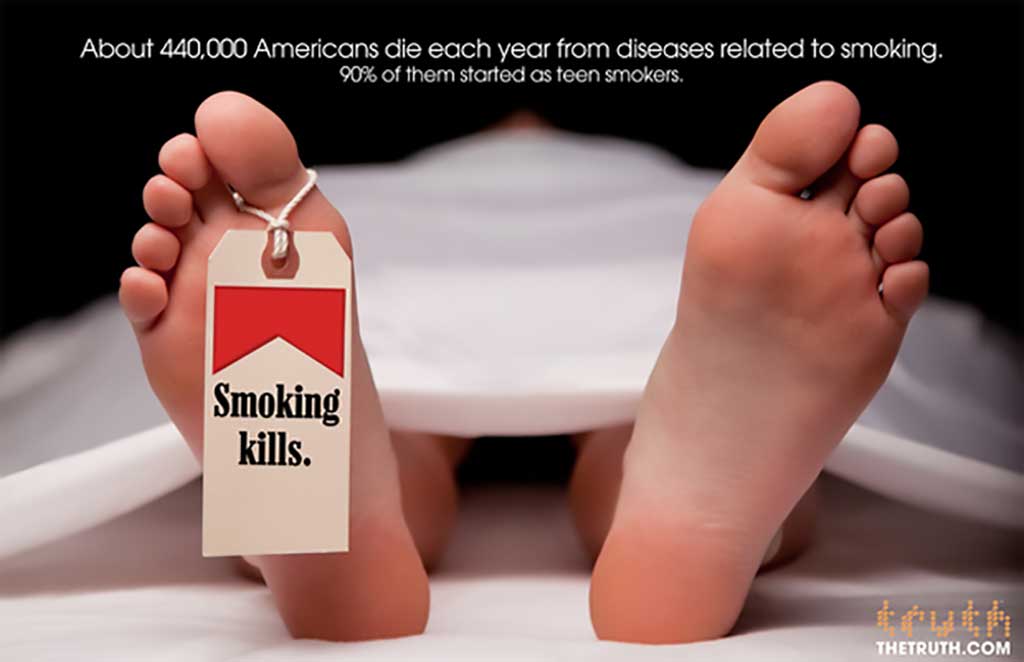 An image of a pair of feat poking out from beneath a white sheet in a morgue. The foot on the left side of the image has a toe tag with the iconic Marlboro Cigarettes red packaging. The toe tag reads 'Smoking Kills.' The text at the top of the advertisement reads ' About 440,000 Americans die each year from diseases related to smoking. 90% of them started as teen smokers.'