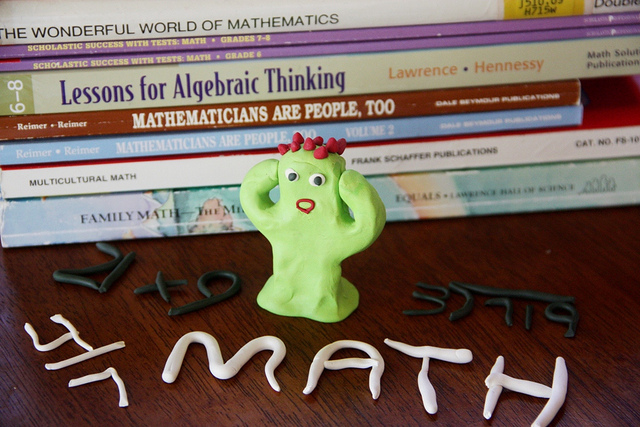 Green figure made out of play-doh, hands to its head, posed in front of a stack of math textbooks. The phrase "math" and simple equations are spelled out in rolled-out play-doh as well.