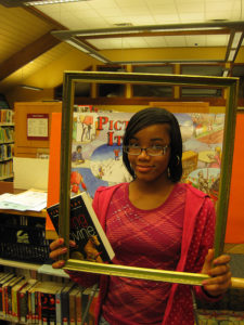 Young woman holding a book and a picture frame in a library, so that she's framed as a reader