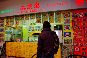 Person with back to camera at counter of Hong Kong Style Wokman's Kitchen. The walls, counter, and menu board are all plastered with menu options.