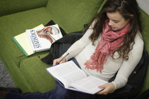 Young woman sitting on a green sofa with a statistics book next to her, reading another book with pencil in hand