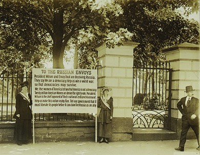 A photograph shows two suffragists standing in front of the White House gate, holding a large sign between them. The text of the sign reads as follows: “President Wilson and Envoy Root are deceiving Russia. They say ‘We are a democracy. Help us win a world war so that democracies may survive.’ We, the women of America tell you that America is not a democracy. Twenty million American Women are denied the right to vote. President Wilson is the chief opponent of their national enfranchisement. Help us make this nation really free. Tell our government that it must liberate its people before it can claim Russia as an ally.”