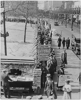 A photograph shows a long line of men waiting on a New York City street for a hot meal. The man at the front of the line holds up a sign that reads, “Line for 1 cent restaurant. 20 meals for 1 cent. Donations invited. Help feed the hungry. 1 cent will feed 20. 1 cent restaurant. 103 W. 43rd St.”