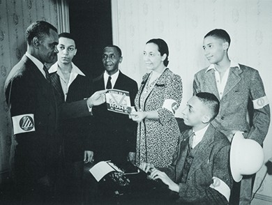 A photograph shows five black men and a black woman participating in the Double V campaign. A young man sits at a typewriter, and the woman hands a man a pamphlet, the cover of which reads “This is a [Double V insignia] Home.” All wear armbands.