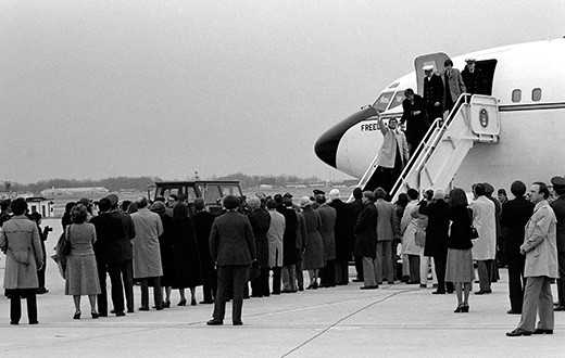 A photograph shows former hostages walking down a flight of steps to exit an official plane; a crowd of people waits for them on the ground.