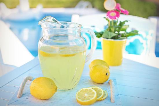 A pitcher of lemonade with lemon slices inside is placed on a blue table near a swimming pool. Whole lemons and lemon slices are beside it, and a yellow flower pot is in the background.
