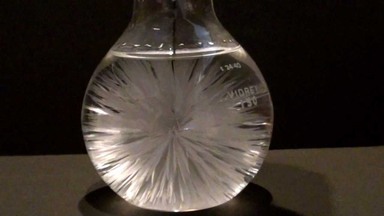 closeup of bulb flask containing liquid with many thin spike crystals radially emerging from the center. 