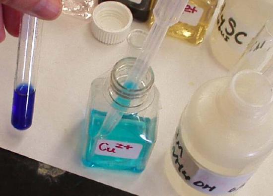 A hand holding a test tube with a liquid that is deep blue in color. On the table is a transparent container of copper solution which pale blue and another container that contains aqueous ammonia. 
