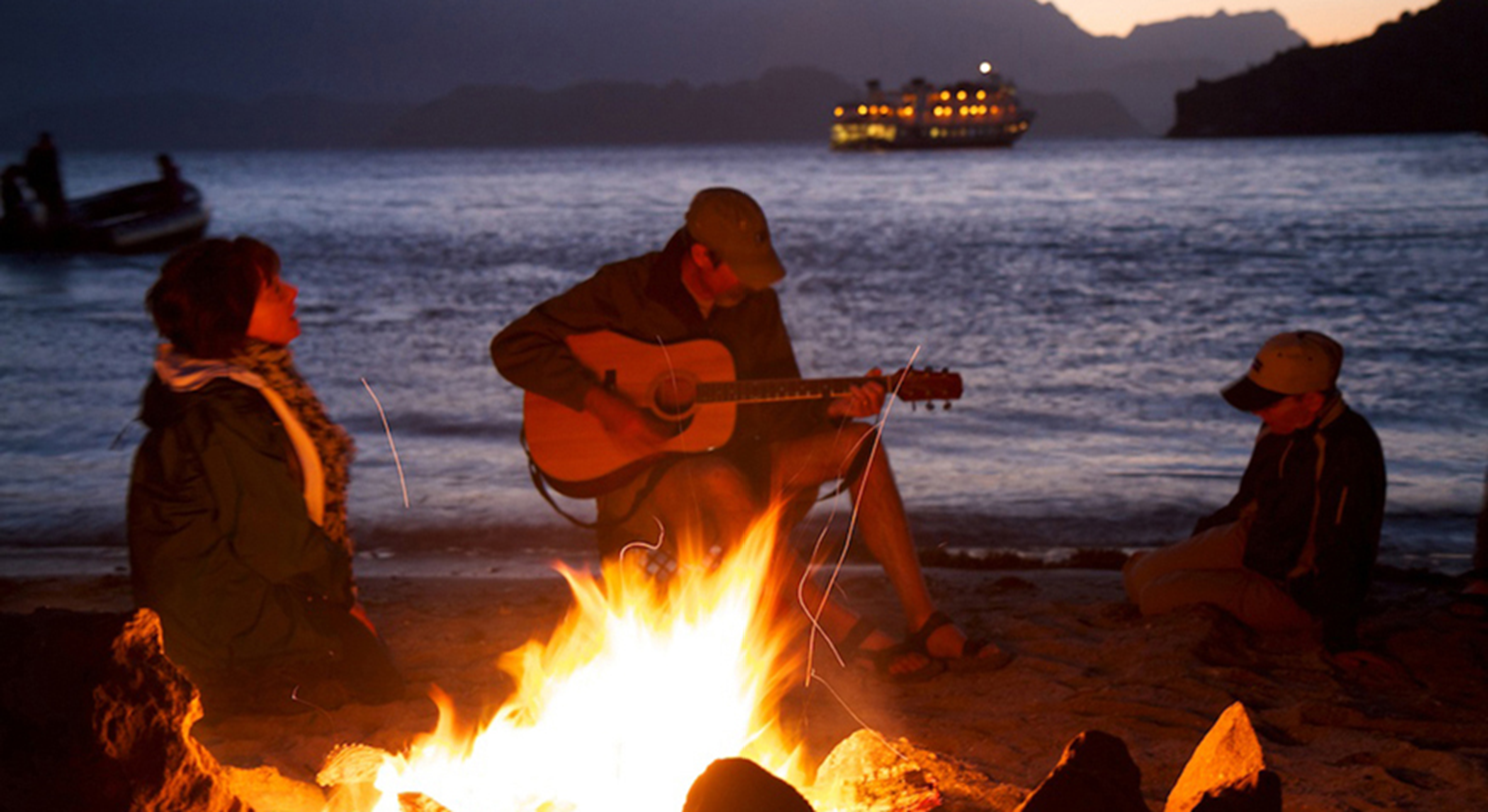 Three people sitting on a beach at night by a fire. One of them is playing a guitar.