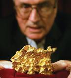 A gold nugget shown in focus in the foreground with an out of focus man behind it.