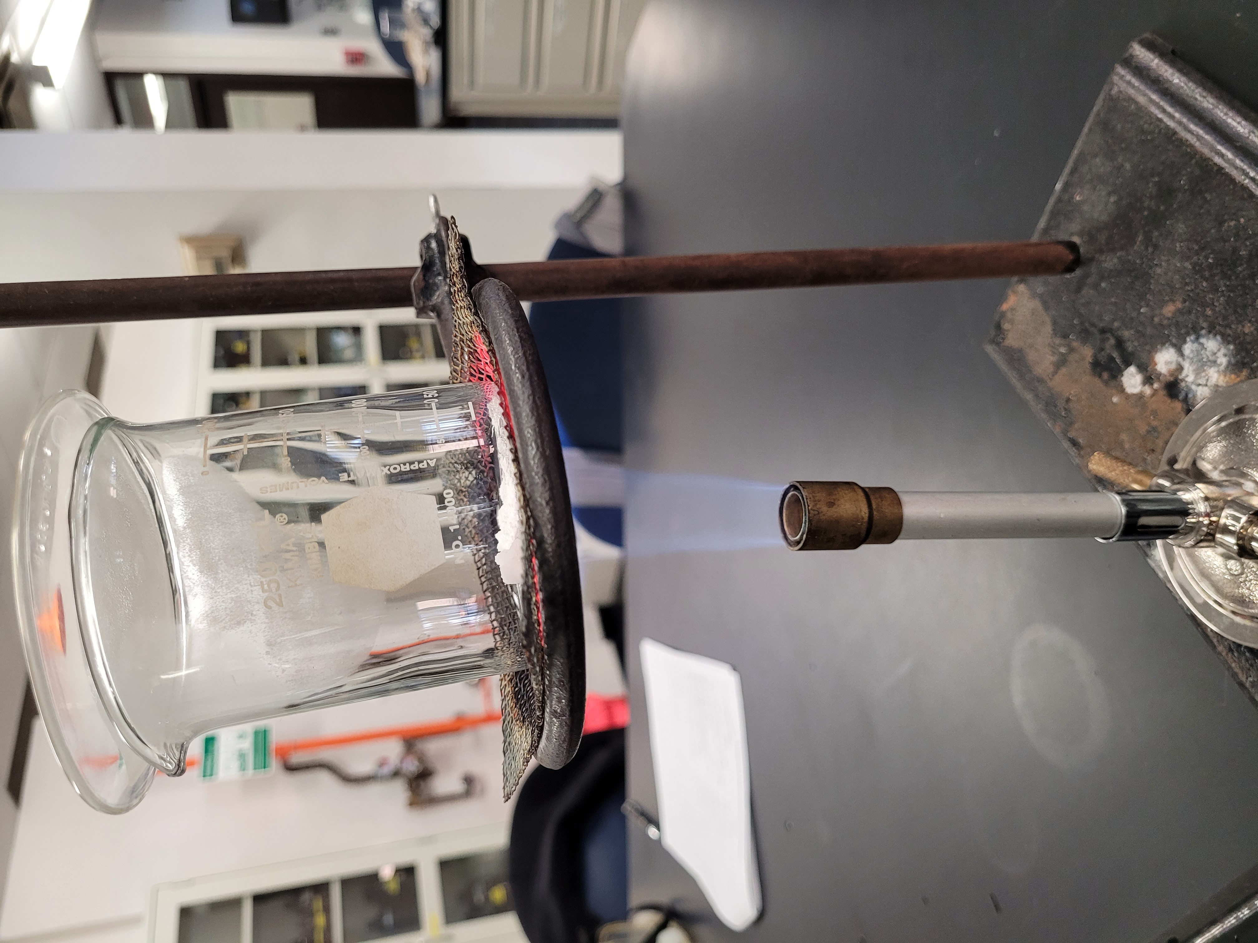 Beaker covered with watch glass containing Alum hydrate is being heated using a Bunsen burner.jpg
