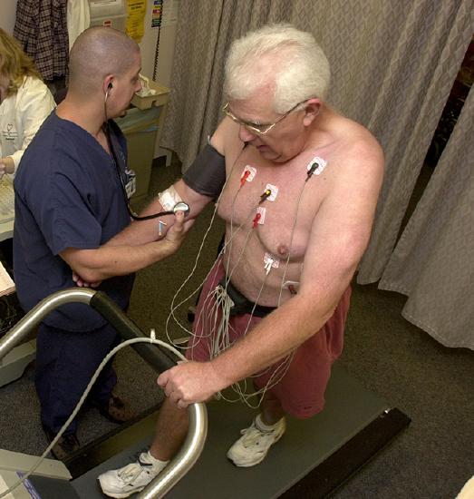 A medical professional is assisting a topless elderly man on a treadmill with sensors and electrical wires connected to his torso.