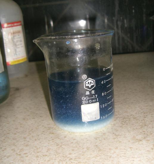 A blue solution is in a glass beaker. White precipitate is forming at the bottom of the beaker.